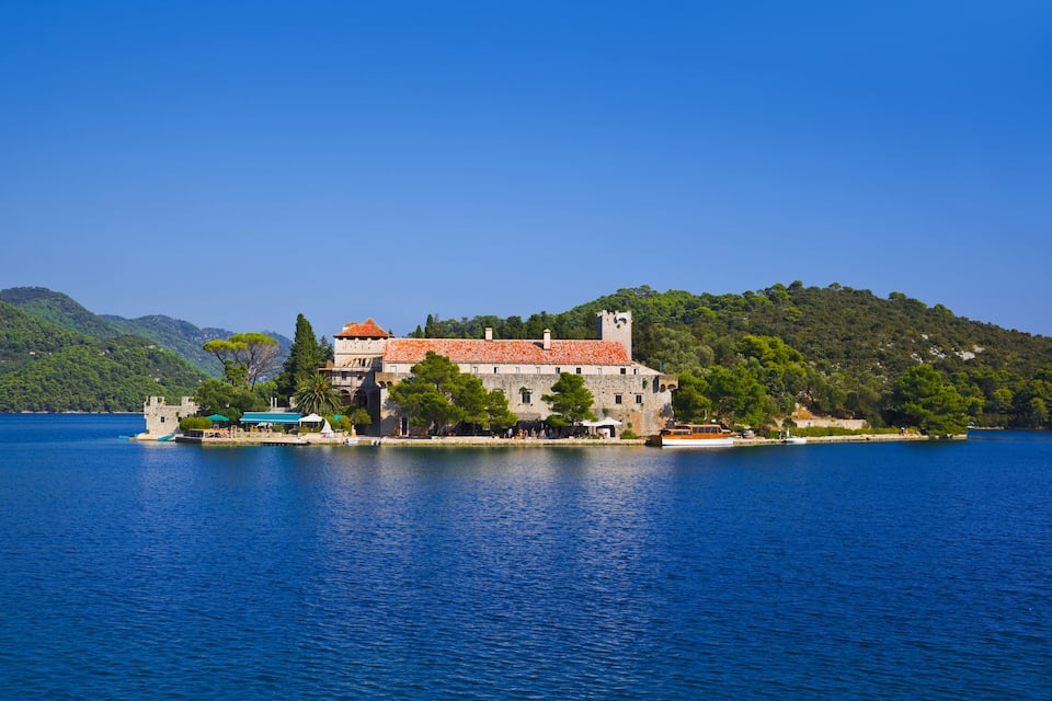 A historic building with a red-tiled roof on a lakeside island, surrounded by lush greenery and hills.