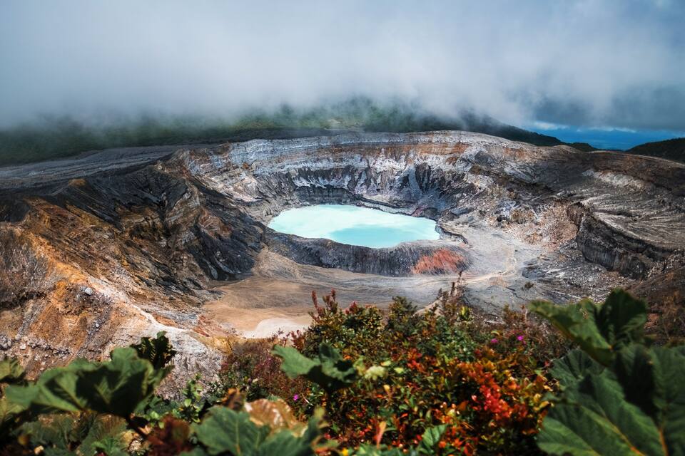 A volcanic crater with a turquoise pool, surrounded by rugged terrain and vegetation.