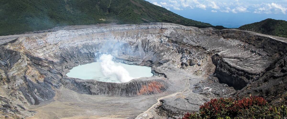 A volcanic crater with a small lake, surrounded by rugged terrain and distant mountains under a partly cloudy sky.