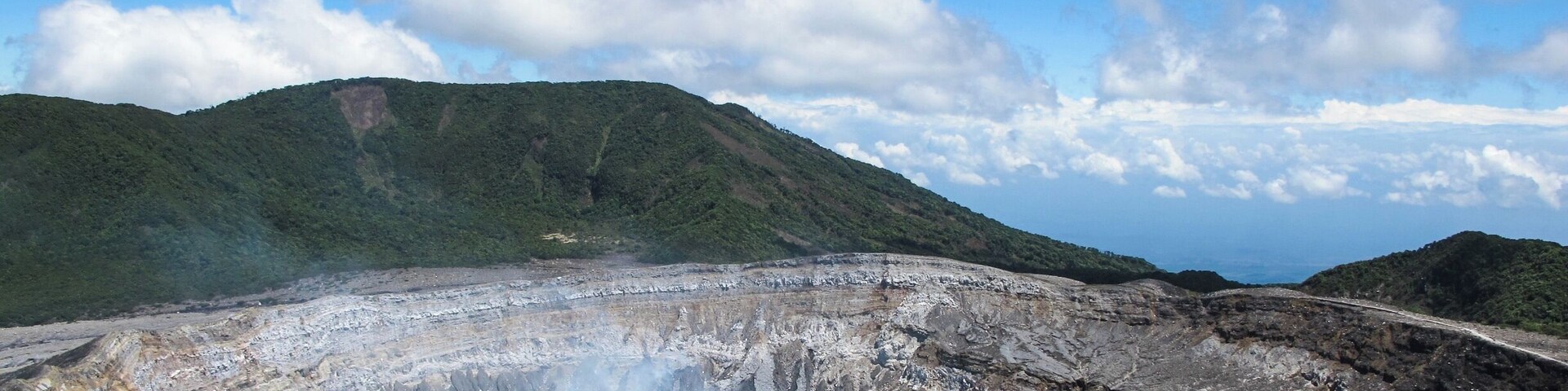 A volcanic crater with a small lake, surrounded by rugged terrain and distant mountains under a partly cloudy sky.