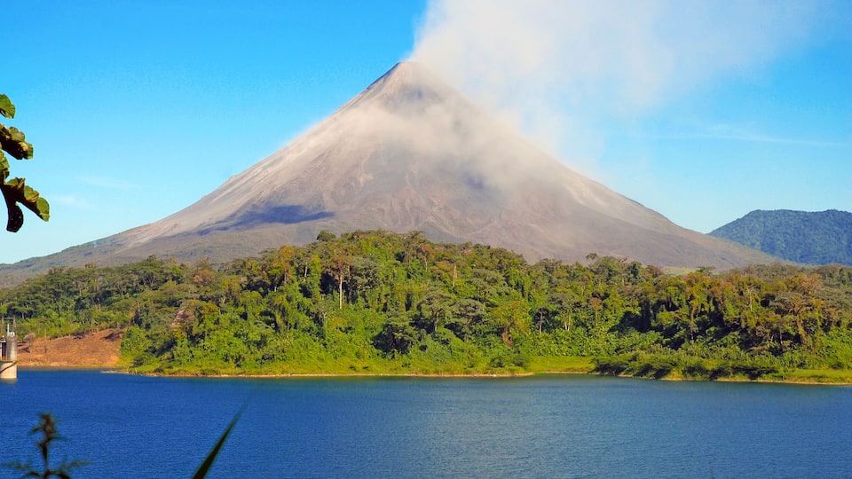 A volcano with a lake in the foreground and forested hills in the background.
