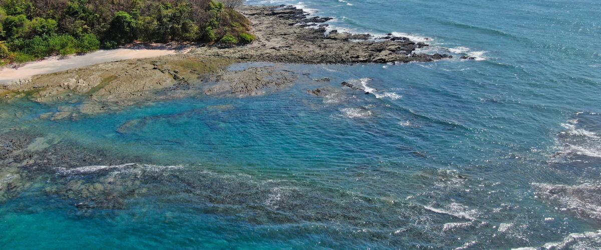 A coastal landscape with a rocky shoreline, clear blue waters, and a forested hill.