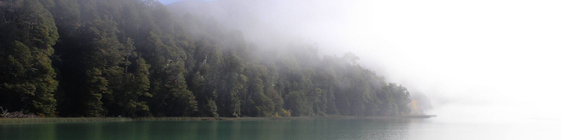 A calm lake surrounded by dense forest and mist-covered mountains.