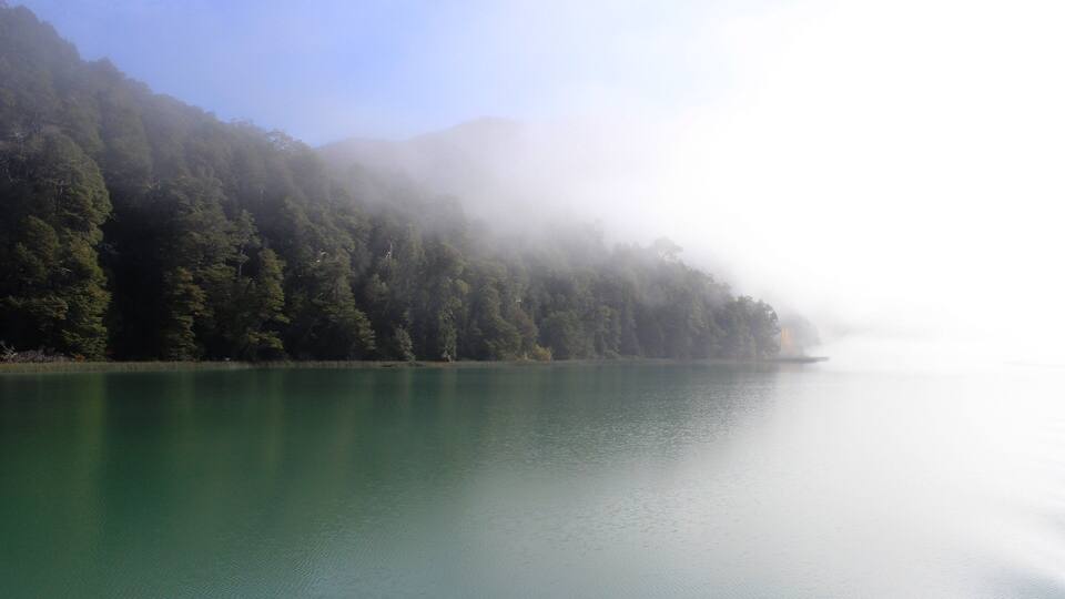 A calm lake surrounded by dense forest and mist-covered mountains.