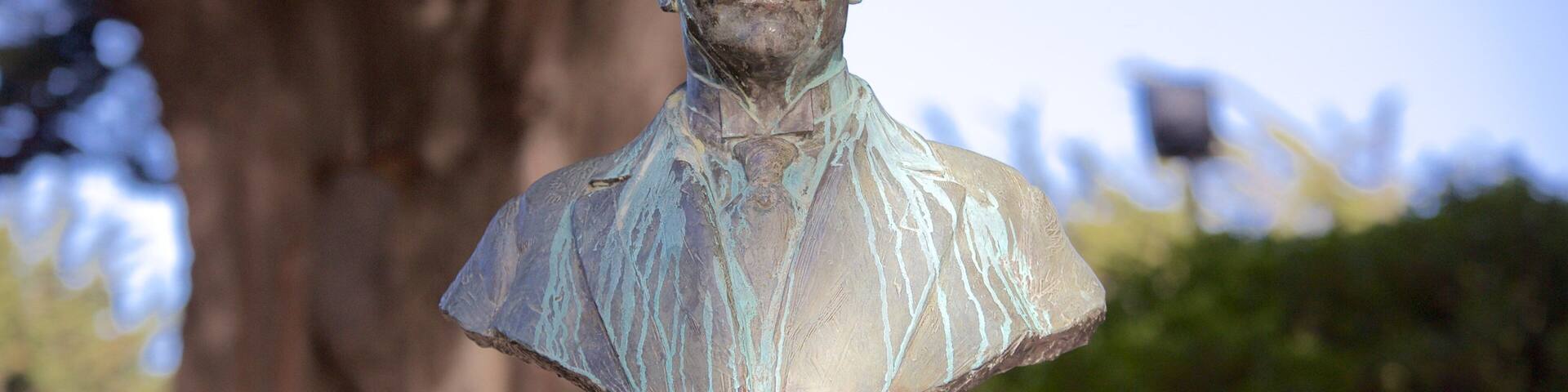 Una escultura de bronce representa un busto de un hombre con traje formal, sobre un fondo de árboles y cielo despejado.