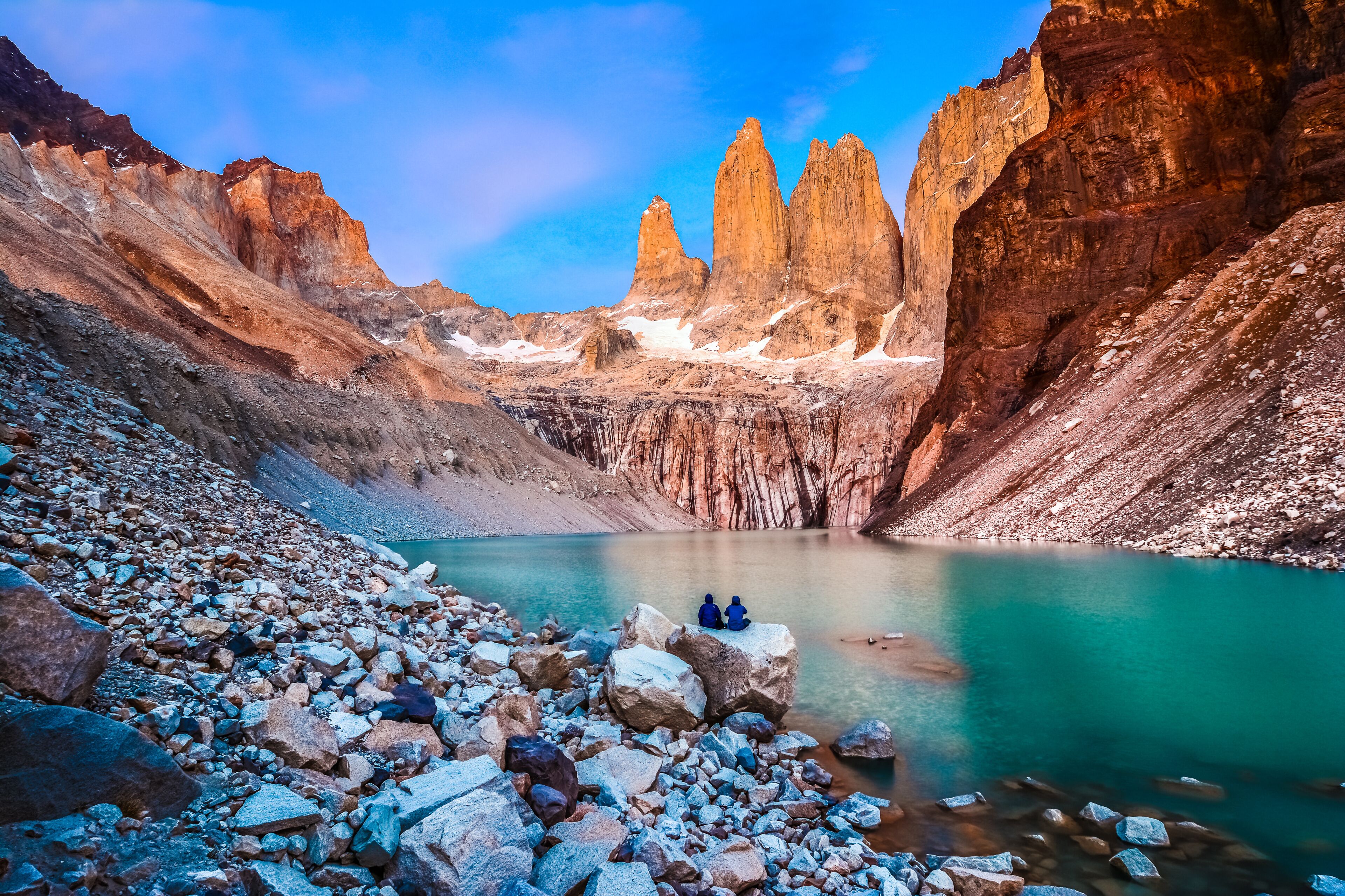 Un paisaje rocoso con un lago turquesa, imponentes acantilados y dos personas sentadas sobre una roca.