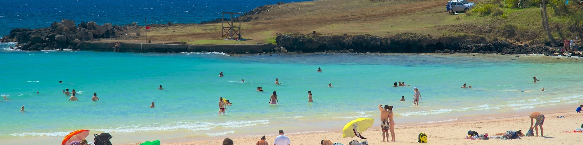 A beach with clear turquoise water, people swimming and sunbathing, and a rocky shoreline.