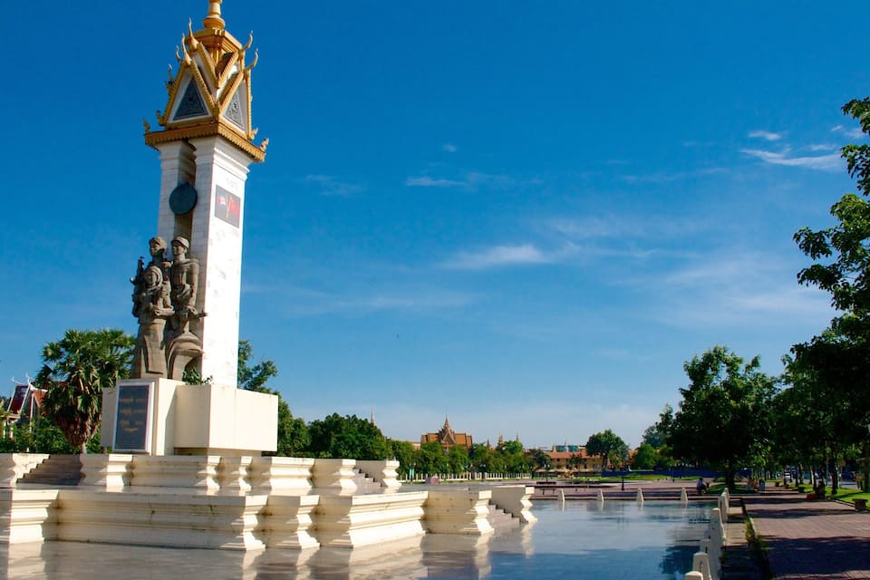 A monument with a golden spire and statues, surrounded by a reflective pool and palm trees.