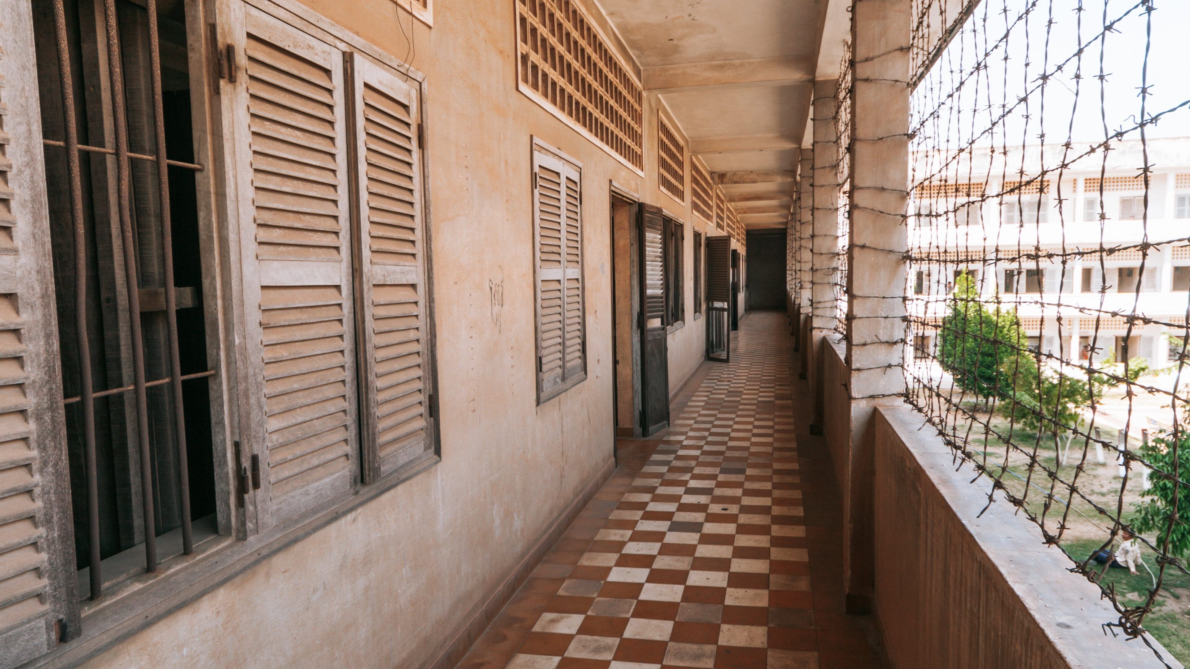 A corridor with checkered flooring, barred windows, and a view of a building with a checkered roof.
