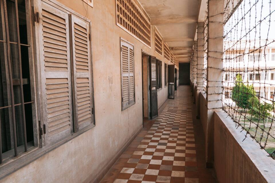 A corridor with checkered flooring, barred windows, and a view of a building with a checkered roof.