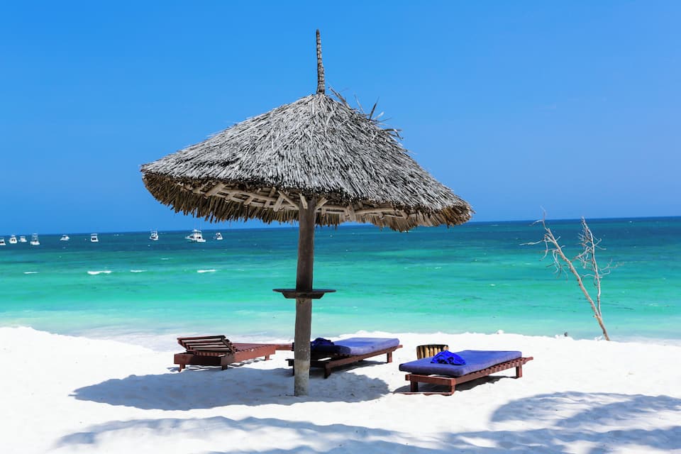 Une plage avec un parasol en chaume, des chaises longues en bois et une mer d’un bleu limpide.