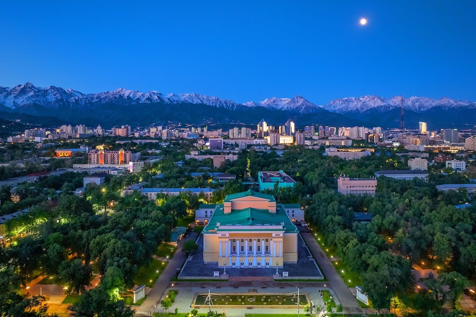 Un paisaje urbano al atardecer con montañas nevadas al fondo y un edificio destacado con techo verde en primer plano.