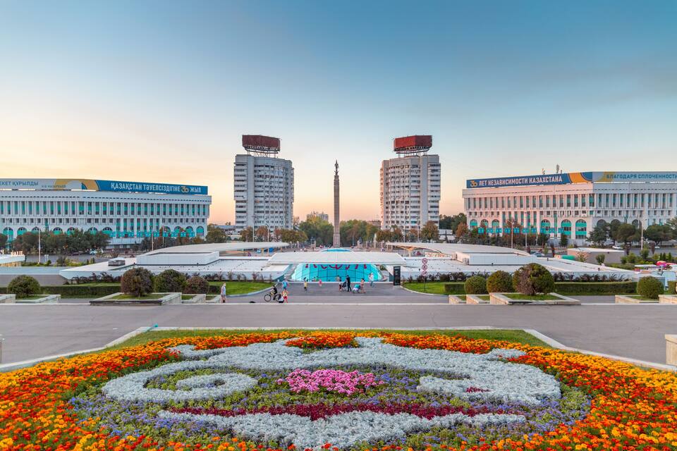 A city square with a flower garden, two tall buildings with banners, and a monument.