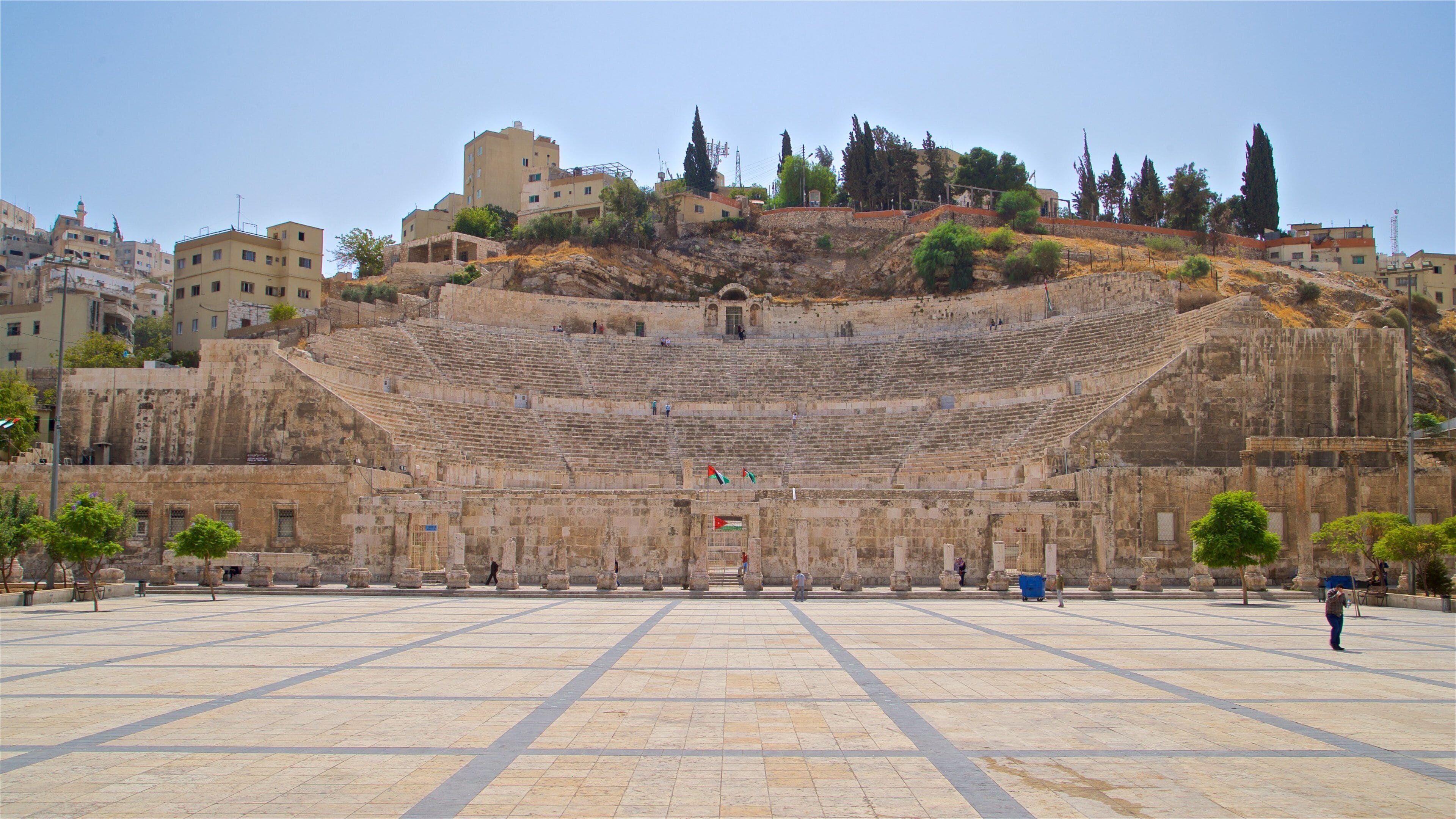 An ancient amphitheater with stone seating and a central stage, surrounded by modern buildings and trees.