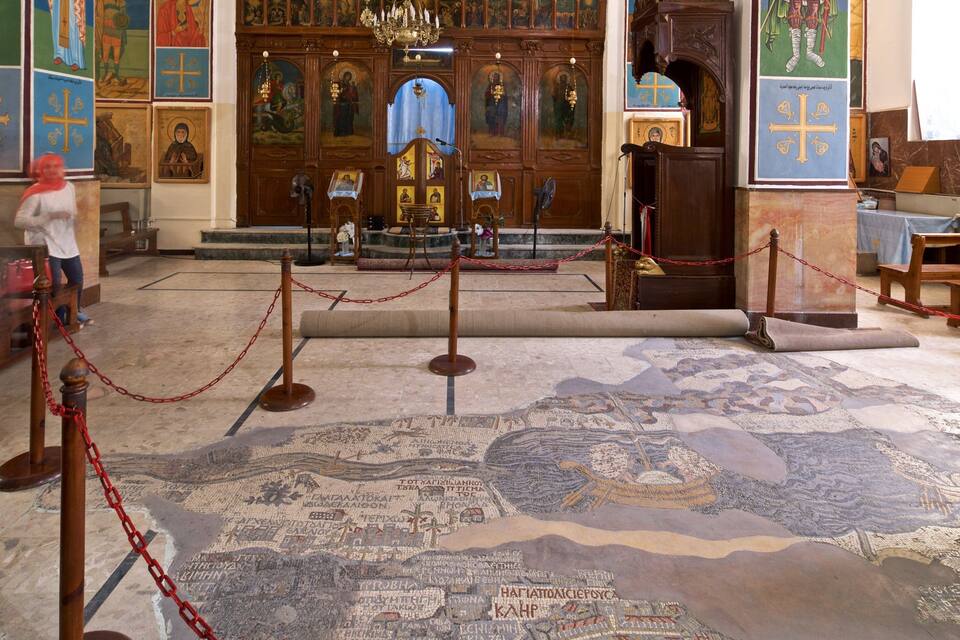 Interior de igreja com ícones religiosos, piso em mosaico e uma pessoa com lenço vermelho na cabeça.