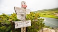 A wooden signpost with multiple directional signs in a mountainous area with a lake in the background.