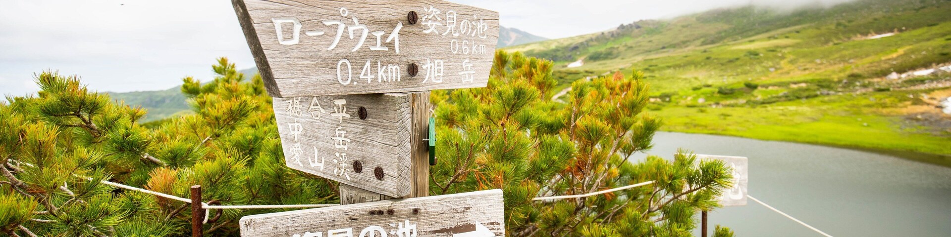 A wooden signpost with multiple directional signs in a mountainous area with a lake in the background.