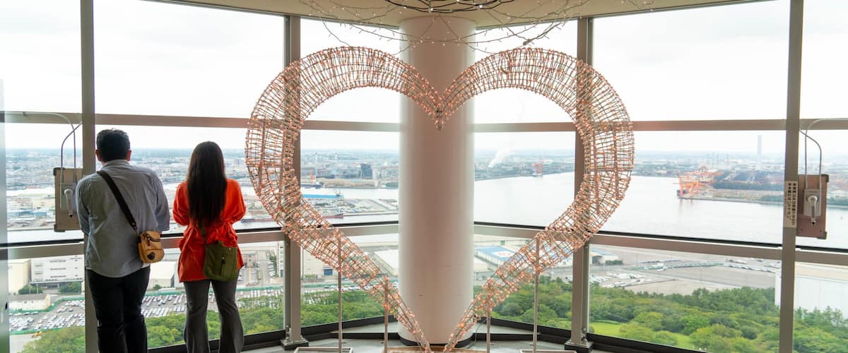 A large heart-shaped sculpture made of wire and lights, viewed from inside a building with large windows overlooking a river and cityscape.