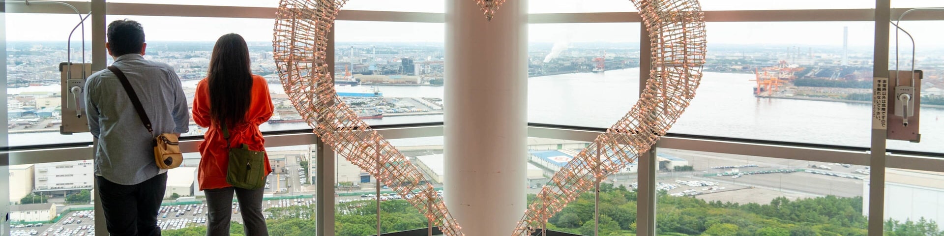 A large heart-shaped sculpture made of wire and lights, viewed from inside a building with large windows overlooking a river and cityscape.