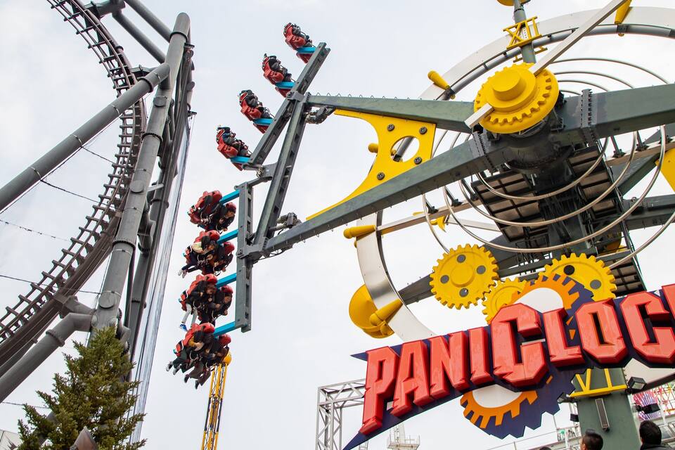 A roller coaster with riders in red harnesses, a large yellow and blue sign reading "PANIC CLOCK", and a Ferris wheel in the background.