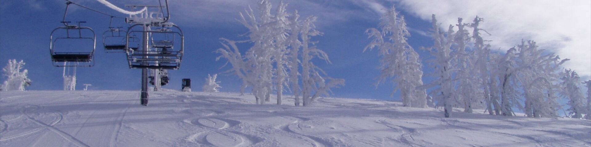 A ski lift chair moving along a snowy slope with tracks, surrounded by snow-covered trees.