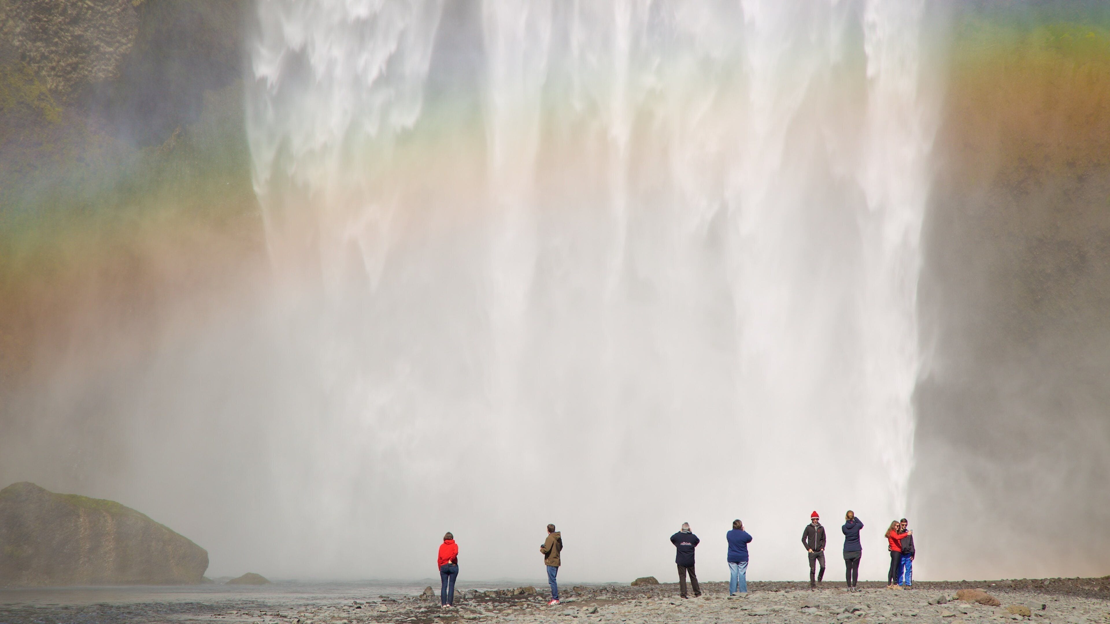 Un grupo de personas está de pie en una costa rocosa, observando una cascada con un arcoíris.