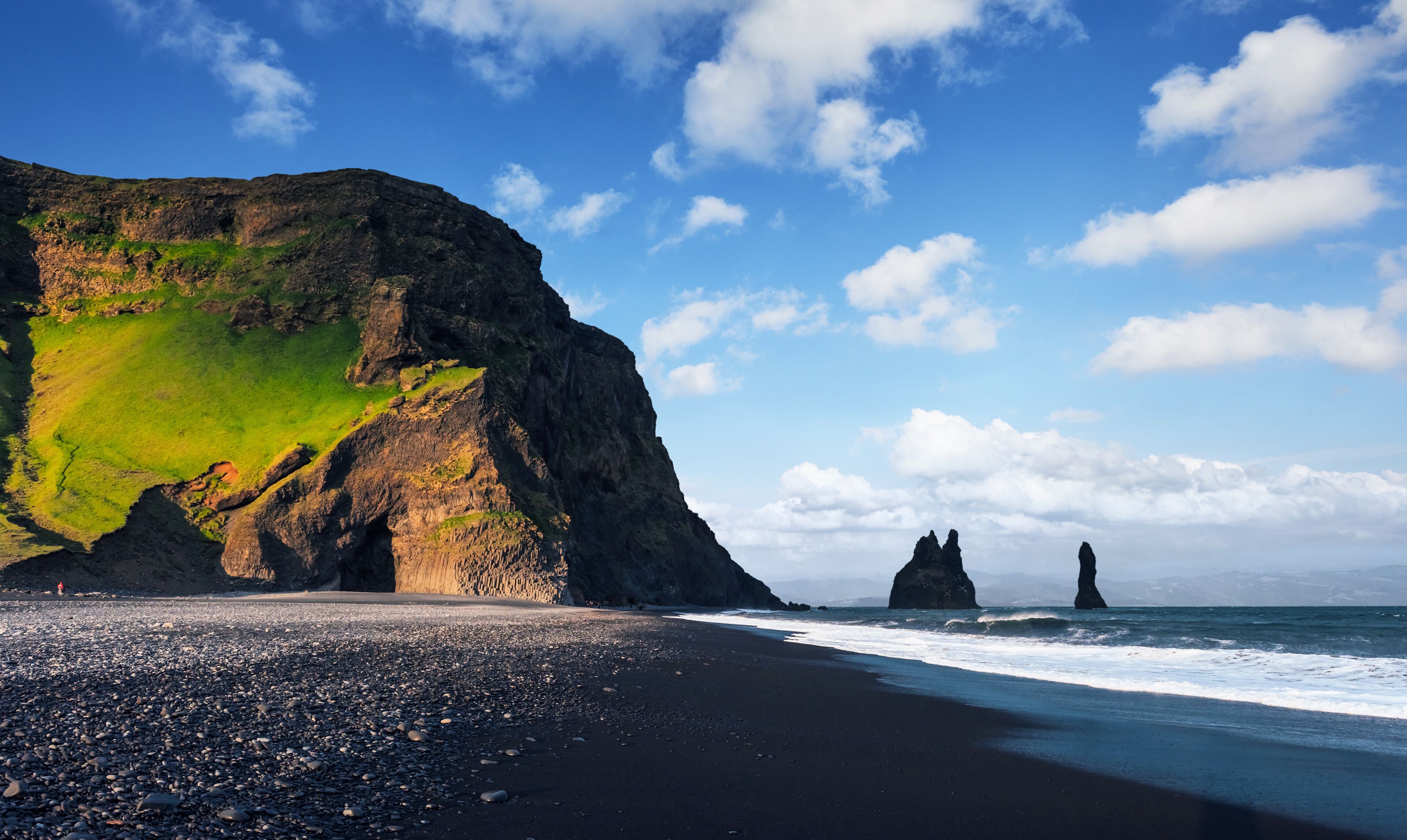 A coastal landscape with a black sand beach, green grassy cliffs, and two prominent rock formations in the sea.