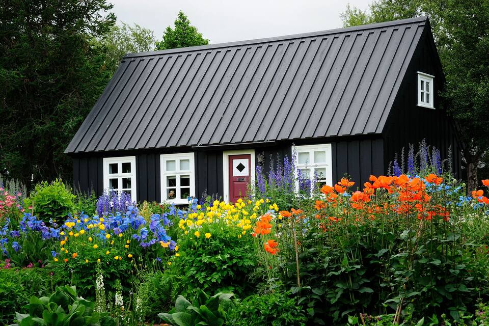 A black wooden house with a sloped roof and white windows, surrounded by a vibrant garden with various colorful flowers.