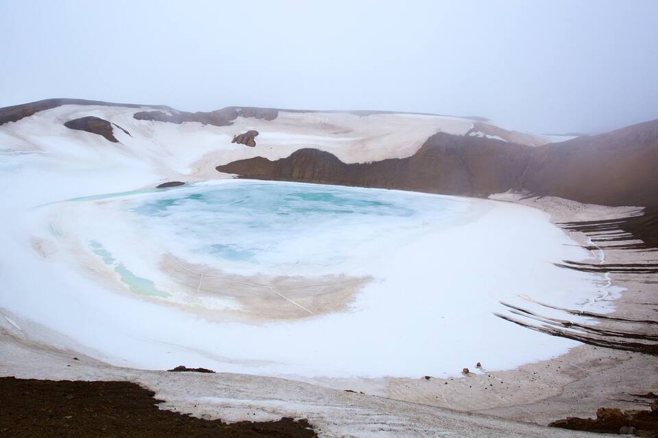 Een geothermisch gebied met een turquoise poel, omringd door sneeuw en rotsachtig terrein.