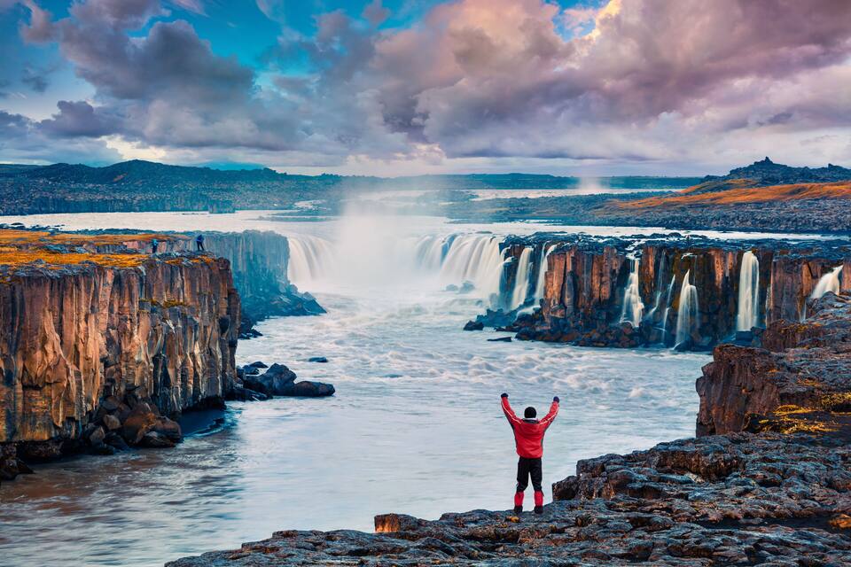 A waterfall cascading over cliffs, a person in red standing on a rocky outcrop, and a vast landscape with distant mountains.