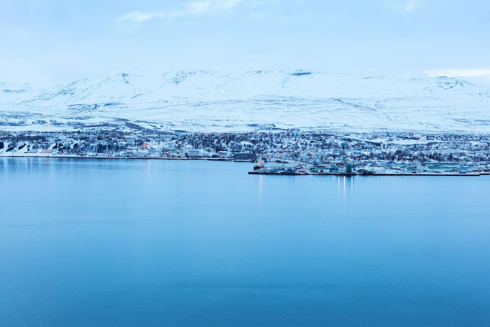 A snow-covered landscape with a calm lake, a small harbor, and a town in the background.