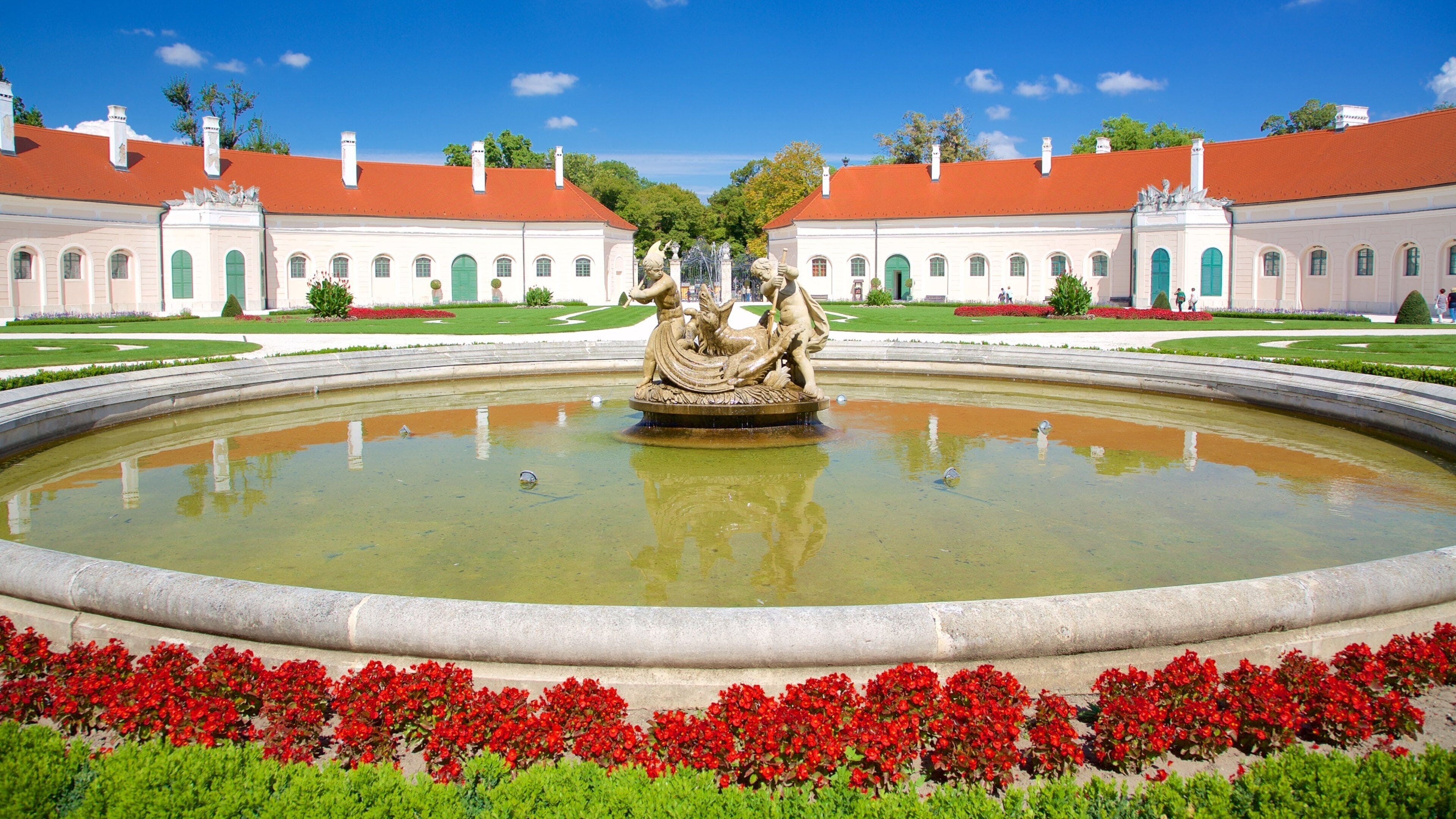 Une fontaine classique ornée de statues, située dans un jardin à la française, entourée de pelouses parfaitement entretenues et de fleurs rouges.