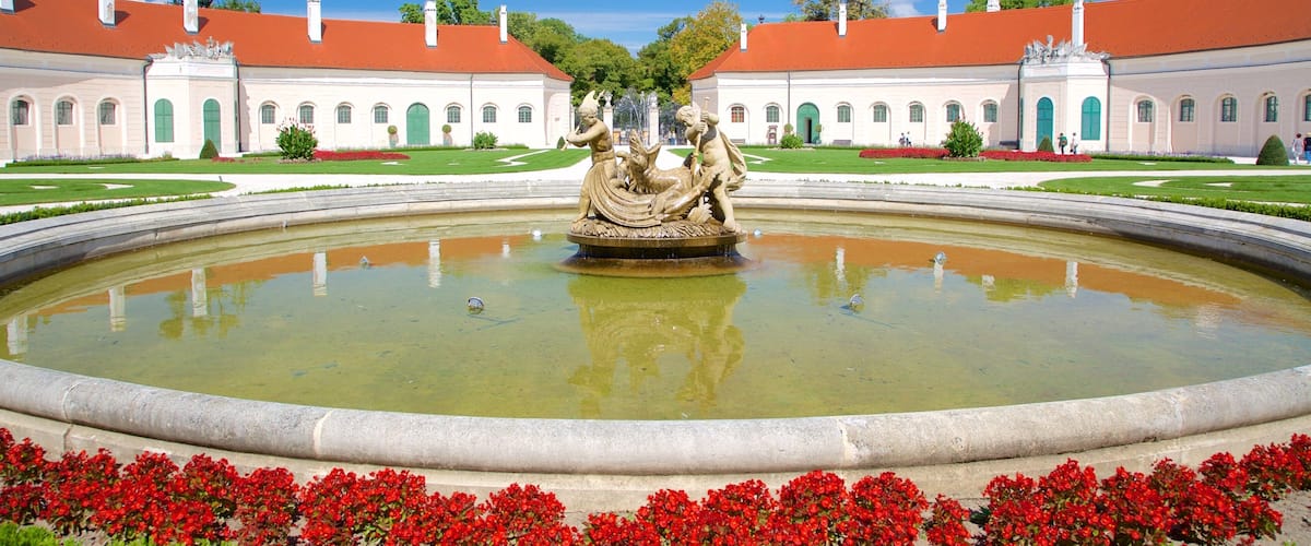 A classical fountain with statues in a formal garden, surrounded by manicured lawns and red flowers.