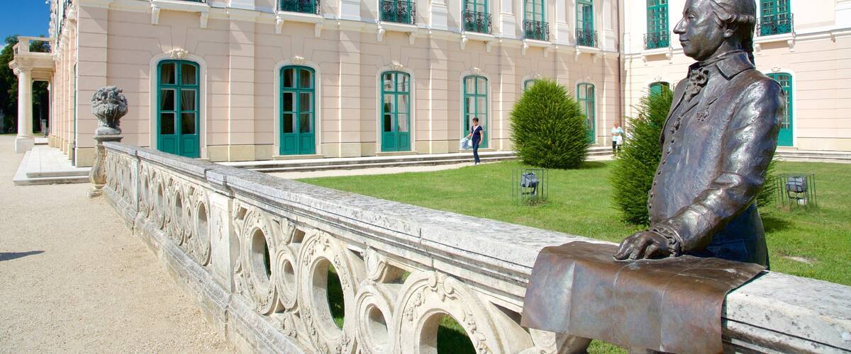 A statue of a man in a suit on a stone balustrade in front of a large building with green windows.