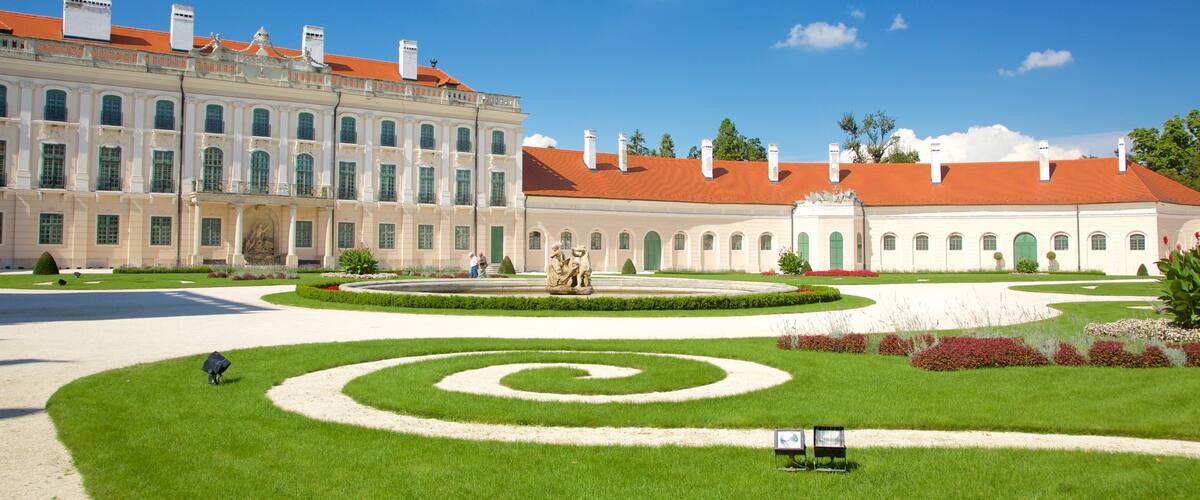 A grand building with a red roof, a large garden with a spiral lawn design, and a statue in the center.