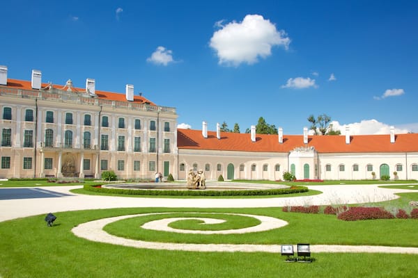 A grand building with a red roof, a large garden with a spiral lawn design, and a statue in the center.