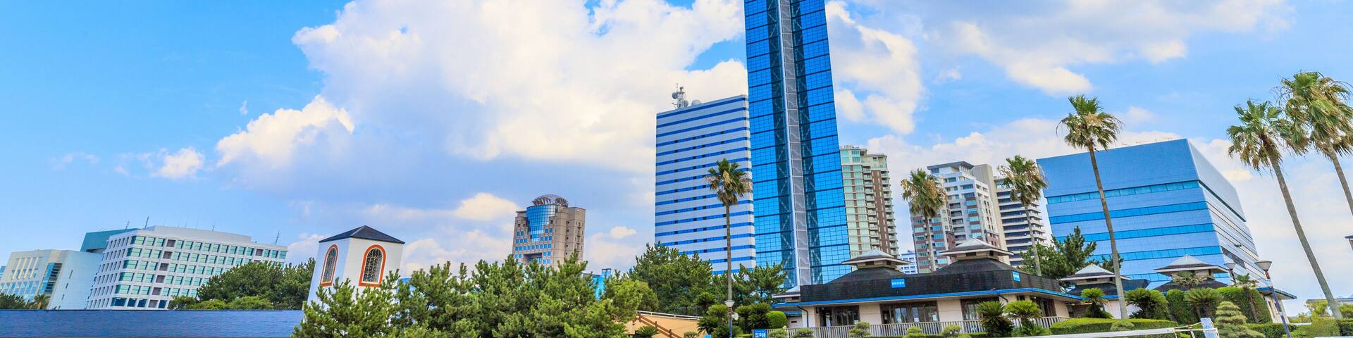 A tall skyscraper with a spire, surrounded by palm trees and other buildings under a blue sky with clouds.
