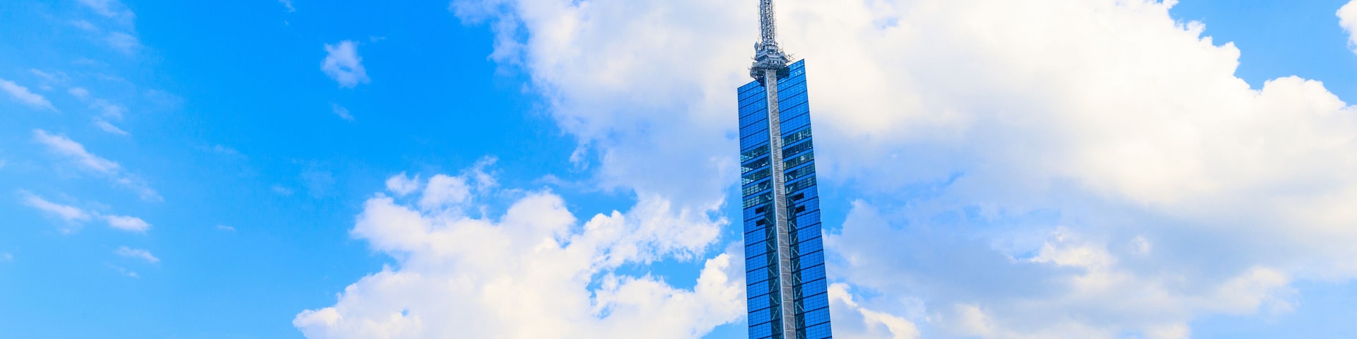 Um arranha-céu alto com um pináculo, rodeado por palmeiras e outros edifícios sob um céu azul com nuvens.