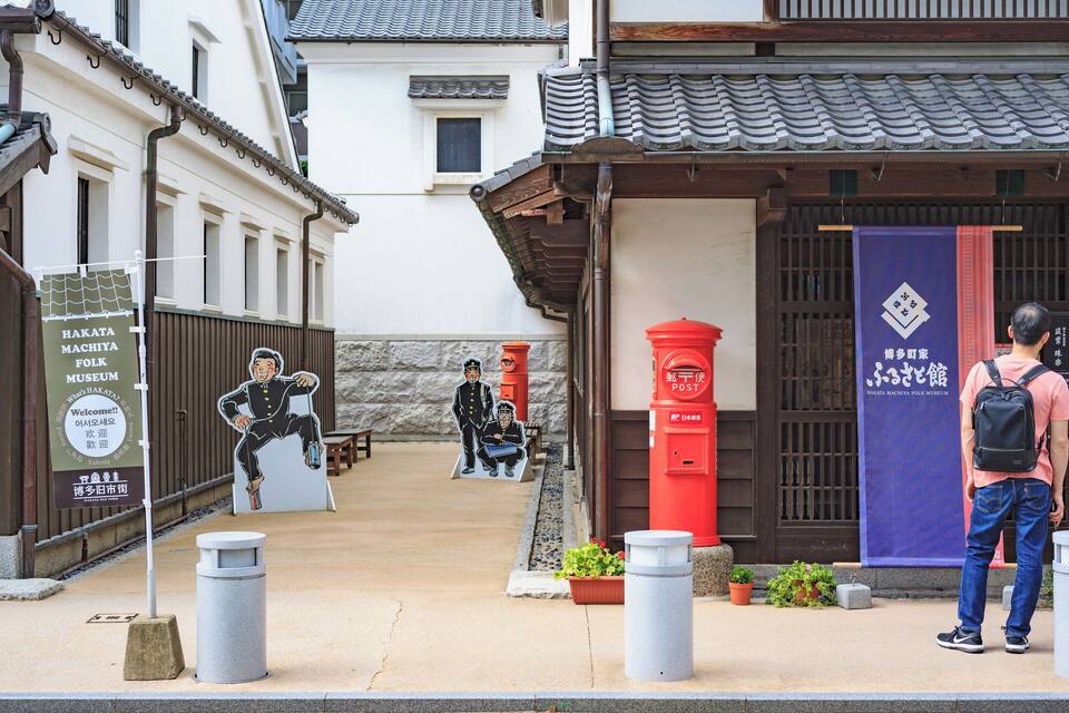 A traditional Japanese building with a red post box and a blue banner. There are two people standing in front of the building.