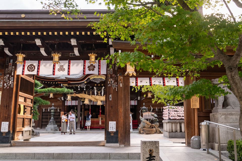 A traditional Japanese temple entrance with wooden architecture, a stone lantern, and cherry blossoms.