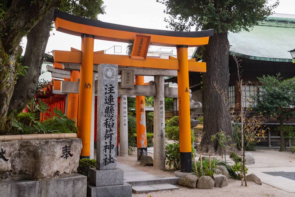 A traditional Japanese torii gate with orange pillars and stone bases, surrounded by trees and rocks.