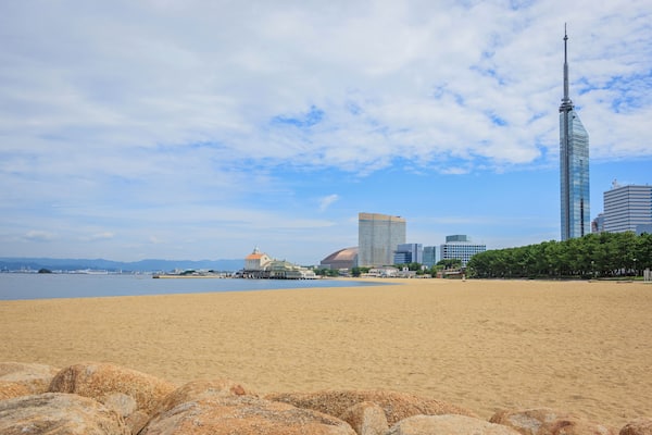 Ein sandiger Strand mit einer großen Felsformation im Vordergrund, ein Gewässer und eine Stadtansicht mit modernen Gebäuden und einem hohen Turm im Hintergrund.