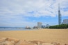 Une plage de sable avec une grande formation rocheuse au premier plan, une étendue d’eau et, au loin, le profil d’une ville avec des bâtiments modernes et une haute tour.