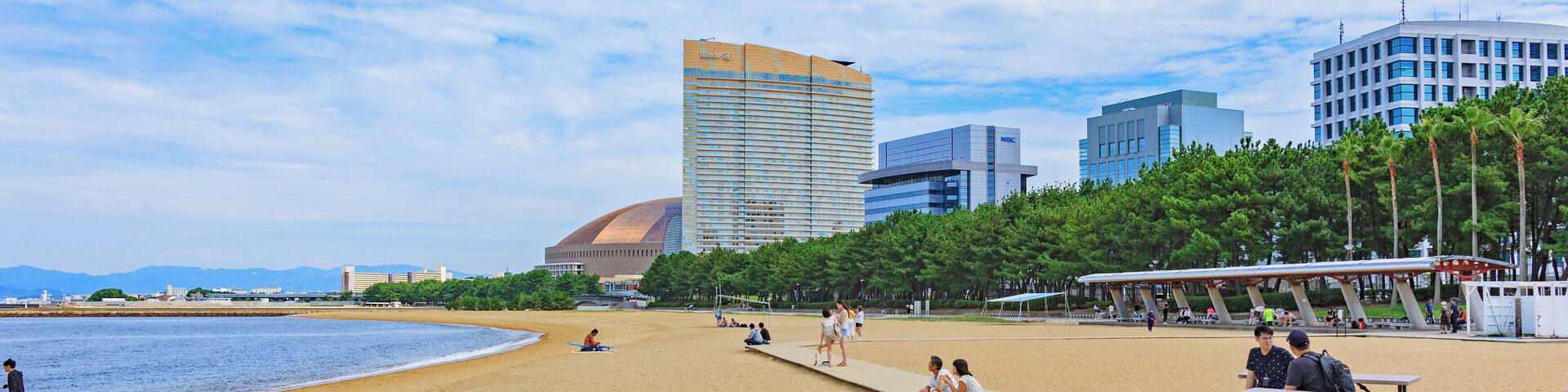A beach with a wooden boardwalk, people sitting on benches, and modern buildings in the background.