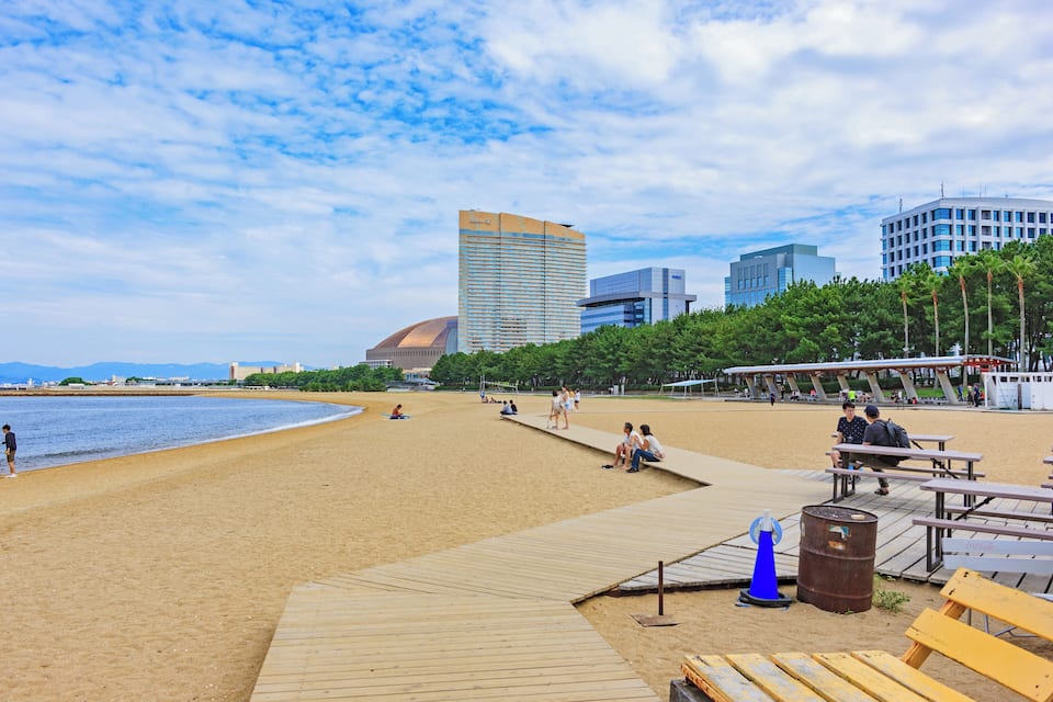 Une plage avec une promenade en bois, des gens assis sur des bancs et, au loin, des bâtiments modernes.