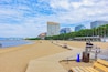 Une plage avec une promenade en bois, des gens assis sur des bancs et, au loin, des bâtiments modernes.