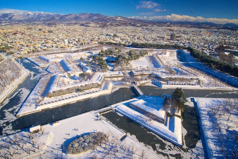 Un parque cubierto de nieve con un estanque central, rodeado de edificios y montañas al fondo.