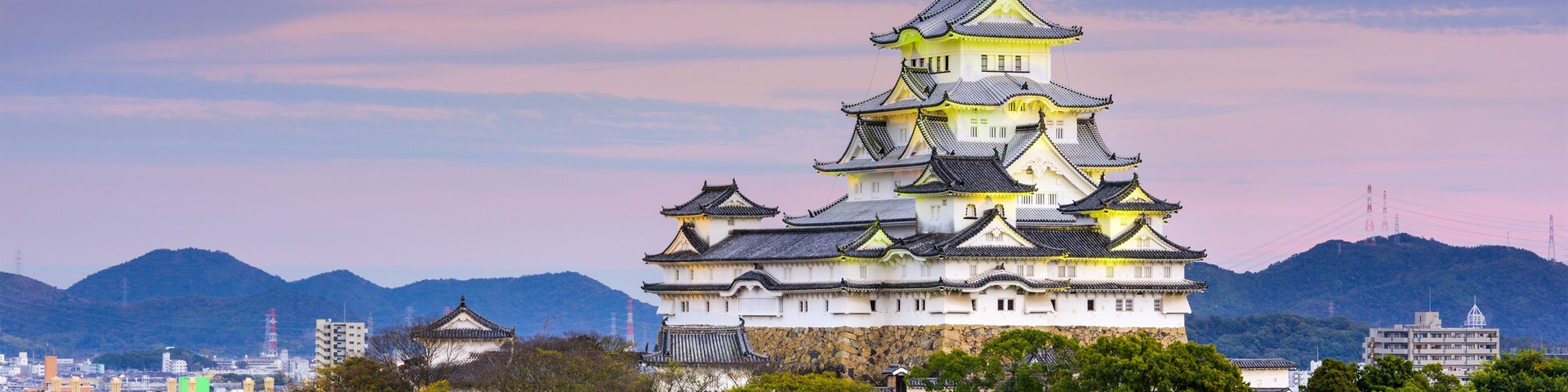 Um castelo japonês tradicional com vários níveis, rodeado por vegetação exuberante e montanhas ao fundo.
