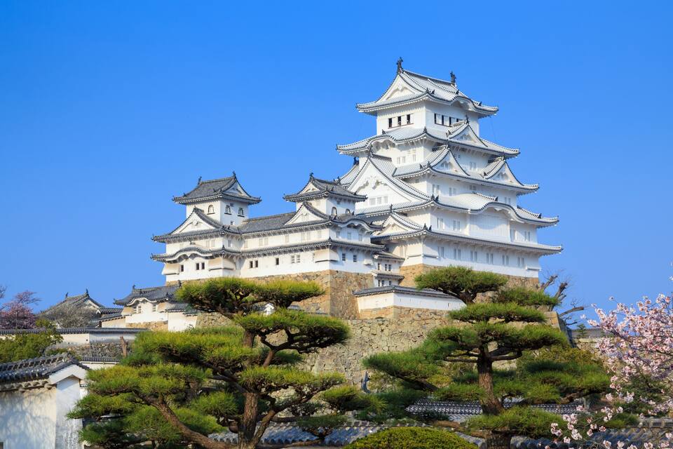 A traditional Japanese castle with multiple tiers, surrounded by lush greenery and flowering trees.