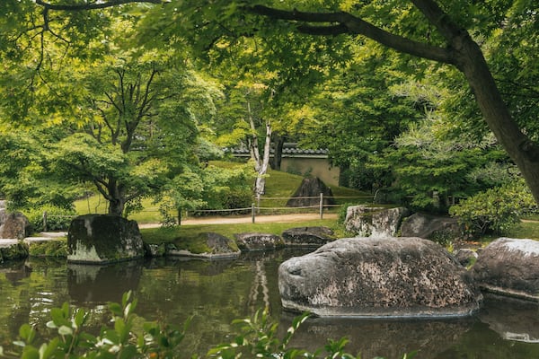 A serene pond with large rocks, lush green trees, and a traditional building in the background.
